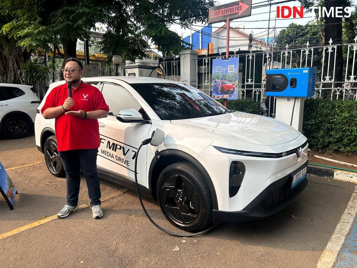 Seorang pria mengenakan kaus merah VinFast berdiri di samping mobil listrik putih VinFast VF MPV 7 yang sedang diisi daya di area parkir.