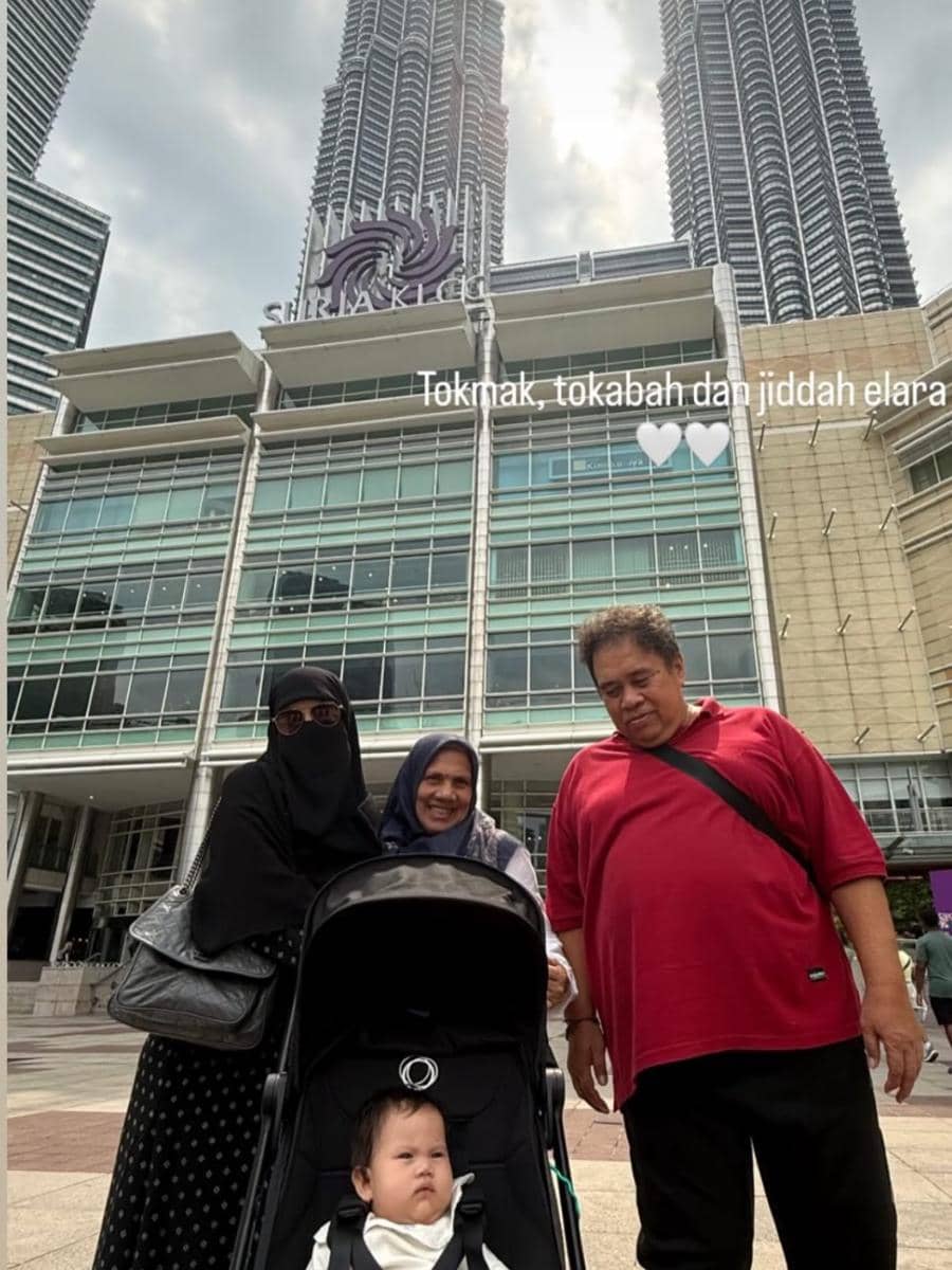 Tiga orang dewasa dan seorang bayi di kereta dorong berpose di depan Menara Kembar Petronas dan Suria KLCC di Kuala Lumpur, Malaysia.