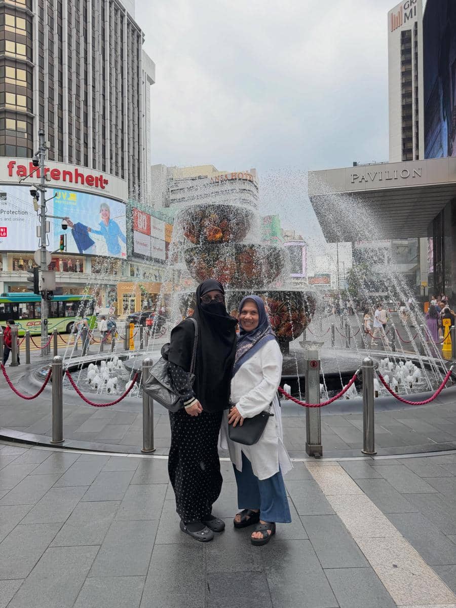 Dua perempuan berpose di depan air mancur besar di kawasan Pavilion Kuala Lumpur, Malaysia, dengan latar gedung dan papan reklame kota.