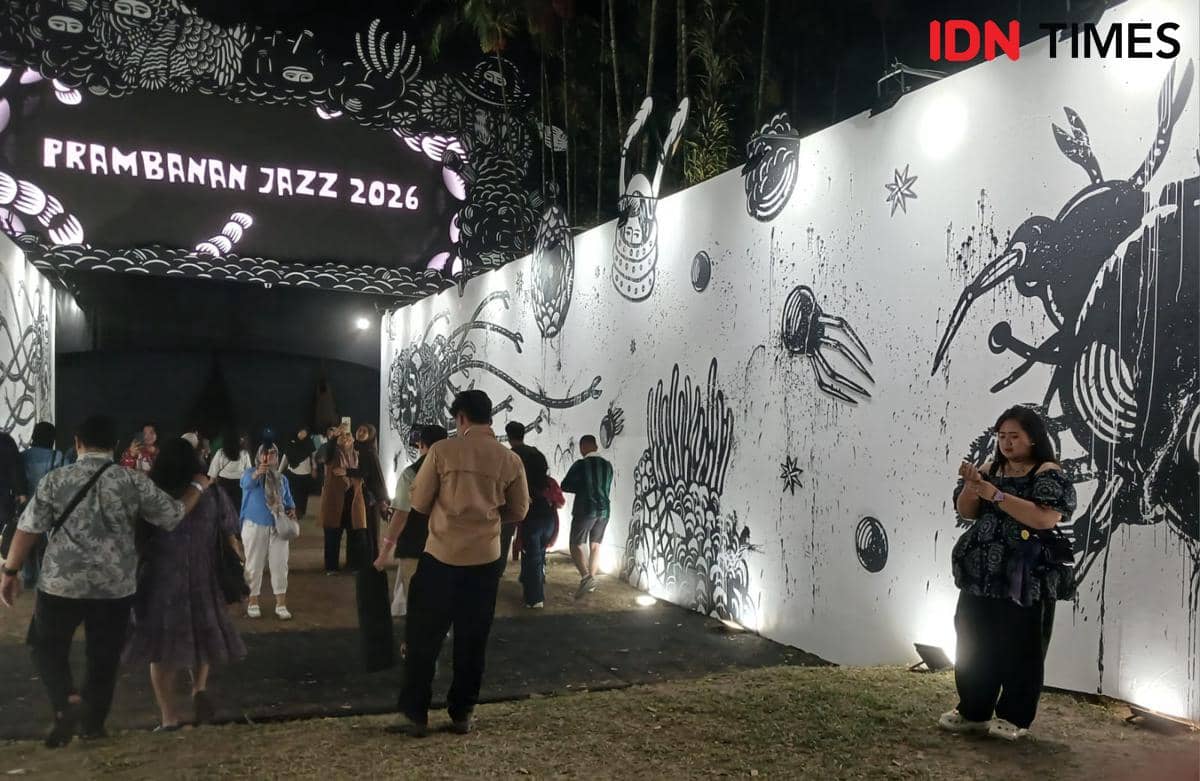 Pengunjung berjalan melewati area masuk Prambanan Jazz Festival 2026 dengan dinding bergambar hitam putih dan pencahayaan malam hari.