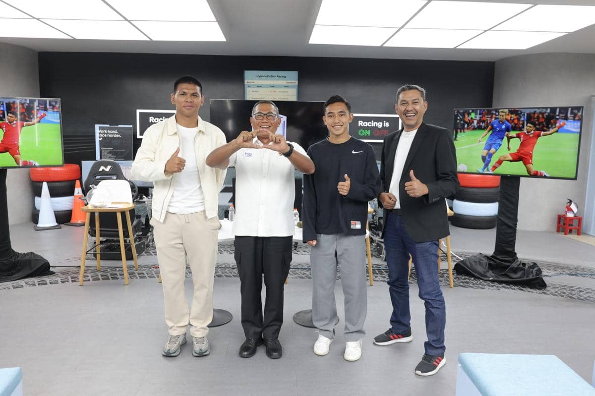 Empat pria berpose sambil tersenyum dan mengacungkan jempol di sebuah ruangan dengan layar menampilkan pertandingan sepak bola di belakang mereka.