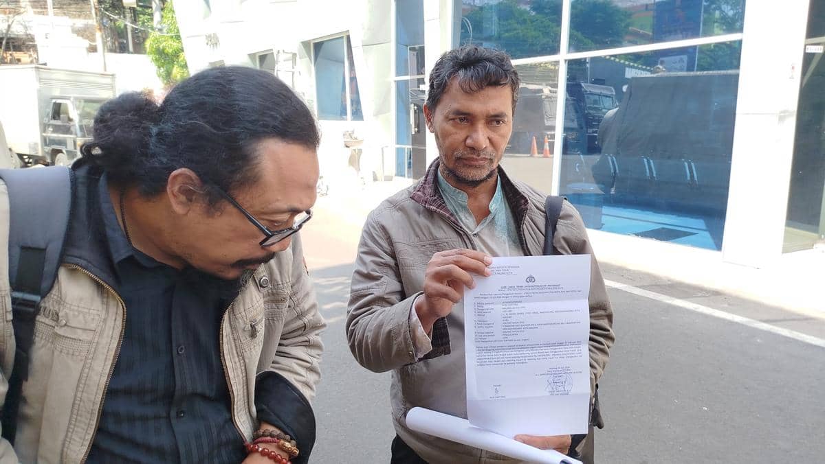 Seorang pria memegang surat laporan polisi di depan gedung Polresta Malang Kota, didampingi seorang pria lain yang mengenakan jaket dan membawa tas.