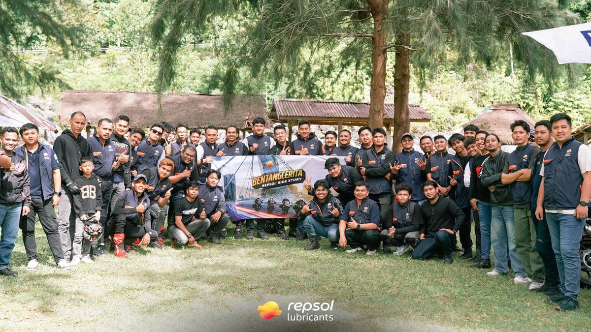 Puluhan peserta komunitas motor berpose bersama sambil memegang spanduk acara Bentang Cerita Repsol Ride Story di area terbuka berhutan.