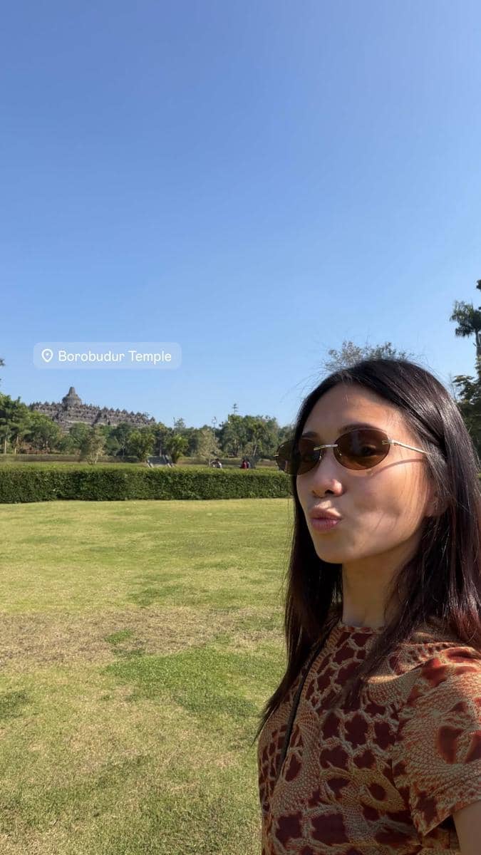 Seorang perempuan berpose dengan latar Candi Borobudur di bawah langit cerah di Yogyakarta, mengenakan kacamata hitam dan pakaian bermotif.