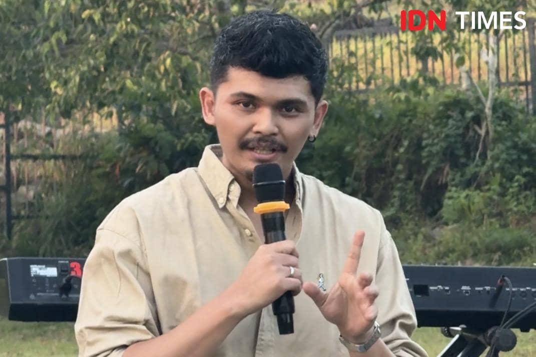 Seorang pria mengenakan kemeja krem memegang mikrofon hitam dengan cincin kuning saat berbicara di luar ruangan dengan latar pepohonan.