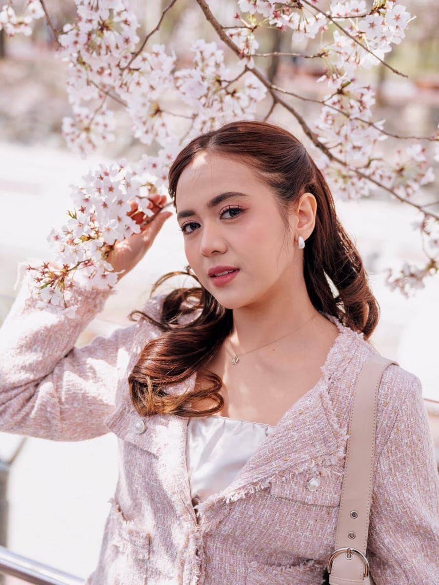 Seorang wanita berpose di bawah cabang pohon sakura bermekaran, mengenakan jaket berwarna lembut dan tas bahu krem.