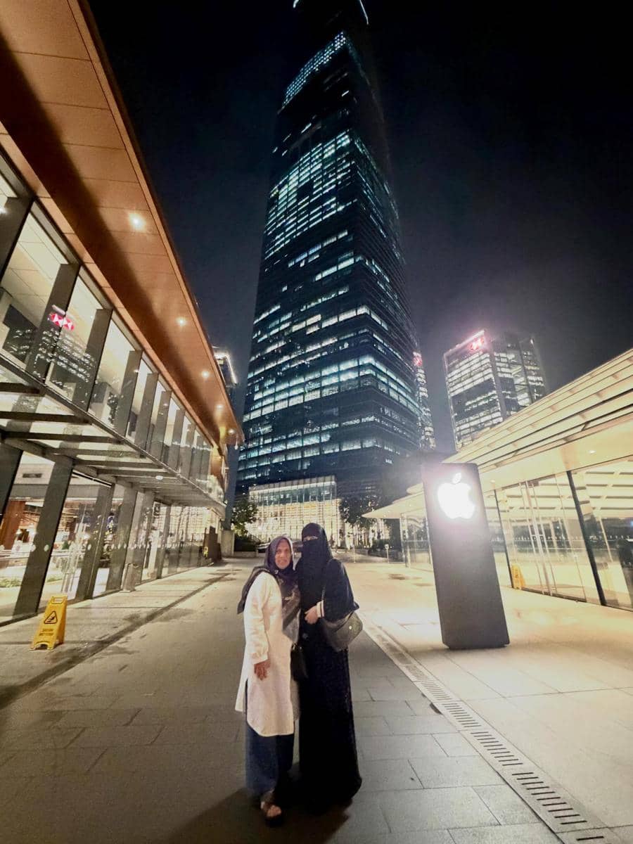 Dua wanita berhijab berpose di malam hari di depan gedung pencakar langit bercahaya di Malaysia dengan suasana kota modern.