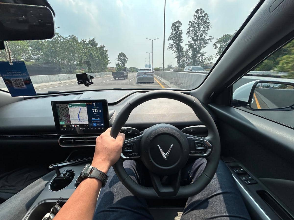Tampilan interior mobil VinFast VF MPV 7 dengan pengemudi memegang setir dan layar navigasi digital aktif di jalan tol.