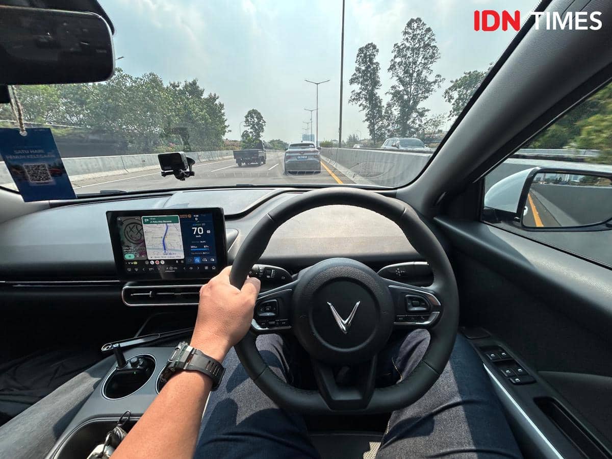 Tampilan interior mobil VinFast VF MPV 7 dengan pengemudi memegang setir dan layar navigasi digital aktif di jalan tol.