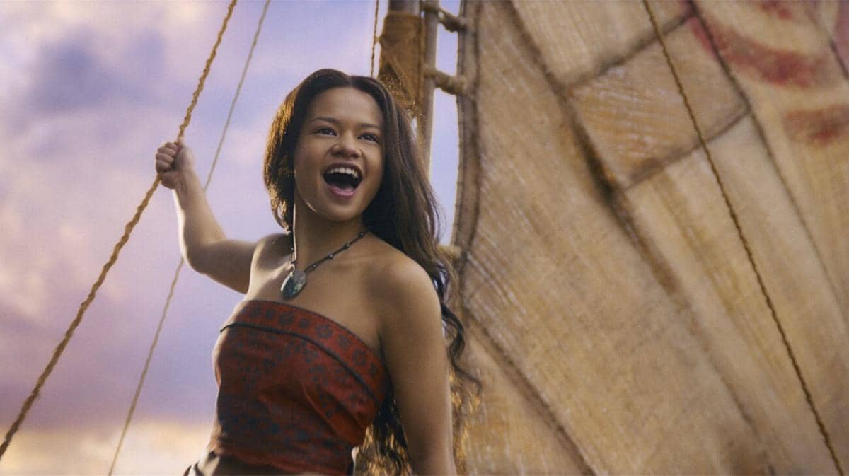 Seorang gadis muda mengenakan pakaian tradisional berdiri di atas perahu layar dengan latar langit senja dalam adegan film Disney's Moana Live Action.