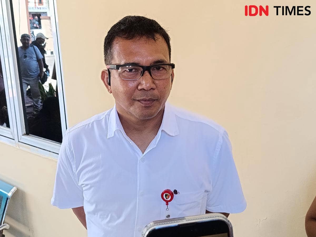 Seorang pria mengenakan kemeja putih dan kacamata berdiri di depan dinding berwarna krem dengan jendela di sampingnya.