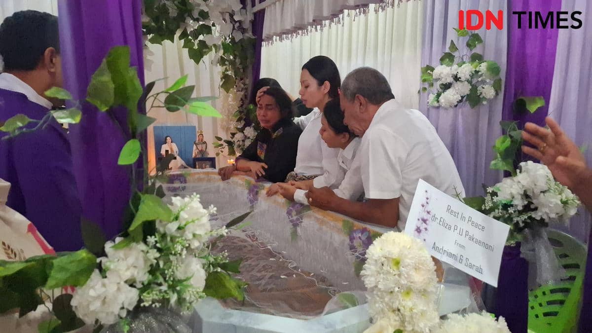 Keluarga berkabung di samping peti jenazah yang dihiasi bunga putih dan ungu pada upacara pemakaman di dalam ruangan.