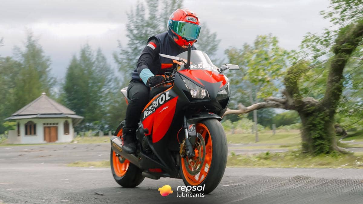Pengendara motor sport dengan livery Repsol mengenakan helm merah dan jaket hitam melaju di jalan dengan latar pepohonan hijau.