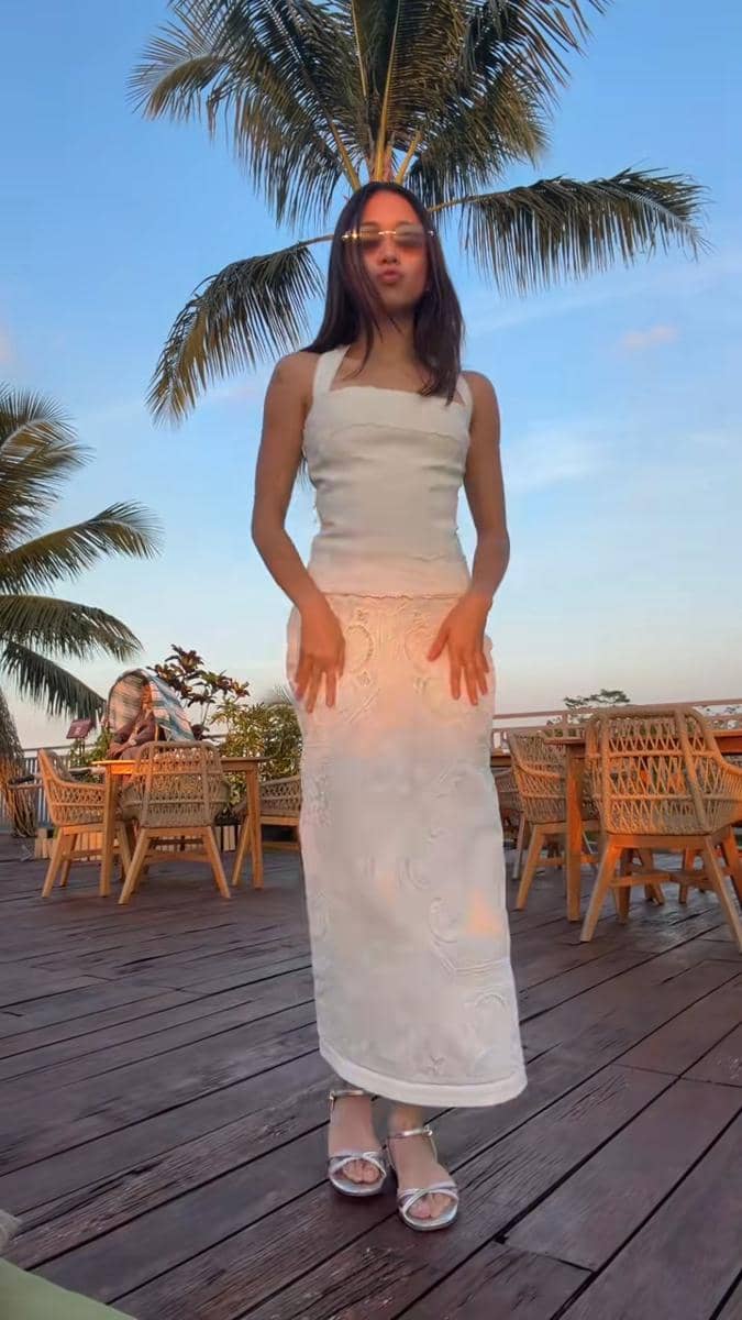 Seorang perempuan mengenakan gaun putih tanpa lengan berpose di area outdoor dengan latar pohon kelapa dan kursi rotan saat matahari terbenam.