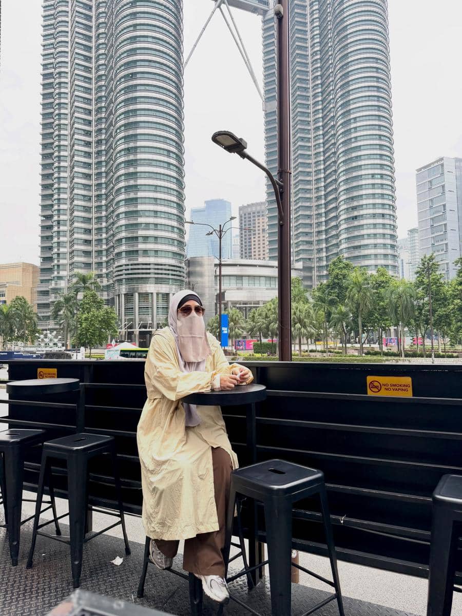 Seorang perempuan berhijab dan berniqab duduk di kafe luar ruangan dengan latar belakang Menara Kembar Petronas di Kuala Lumpur, Malaysia.