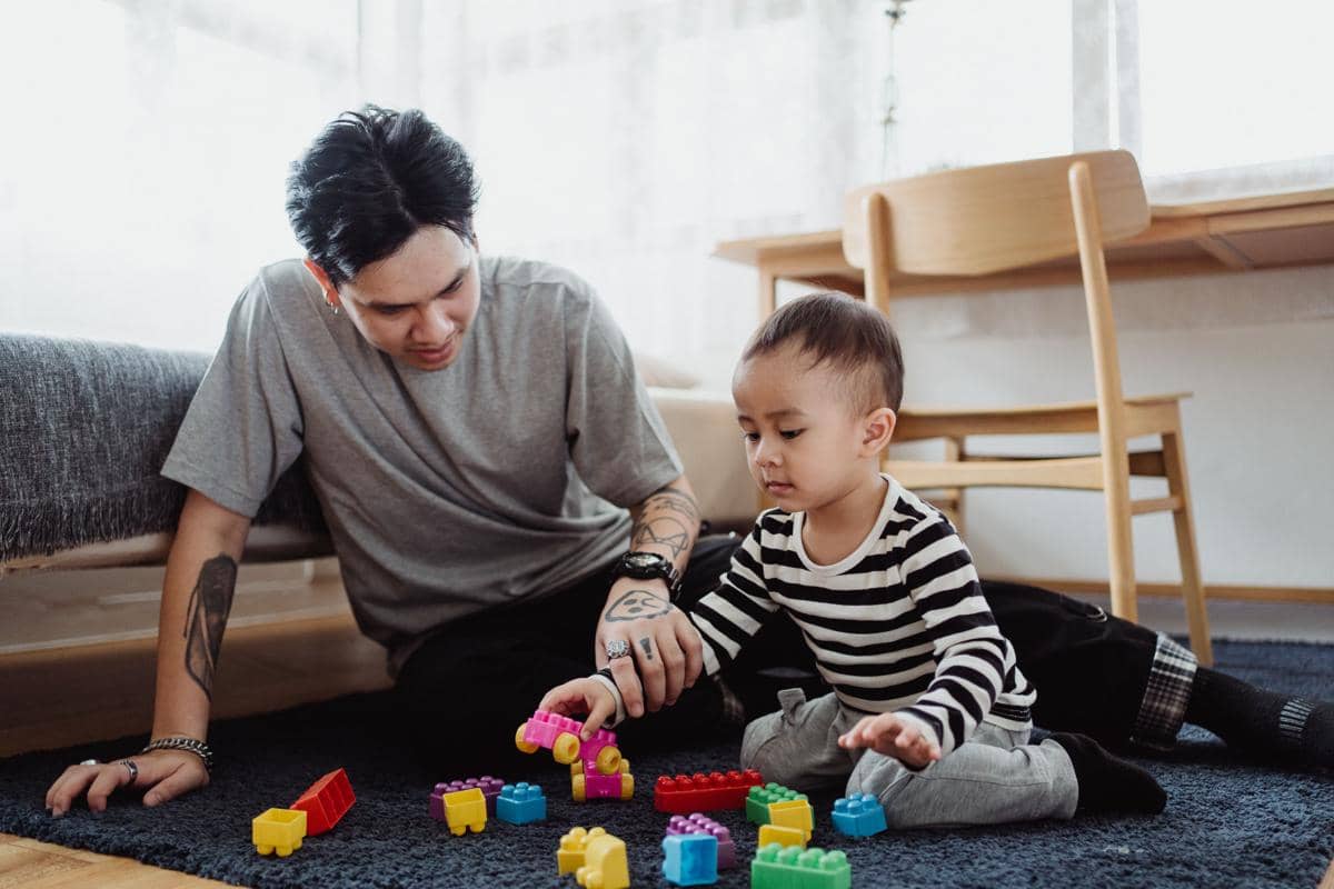 Seorang pria duduk di lantai bersama anak kecil sambil bermain balok warna-warni di ruang tamu yang terang dan nyaman.