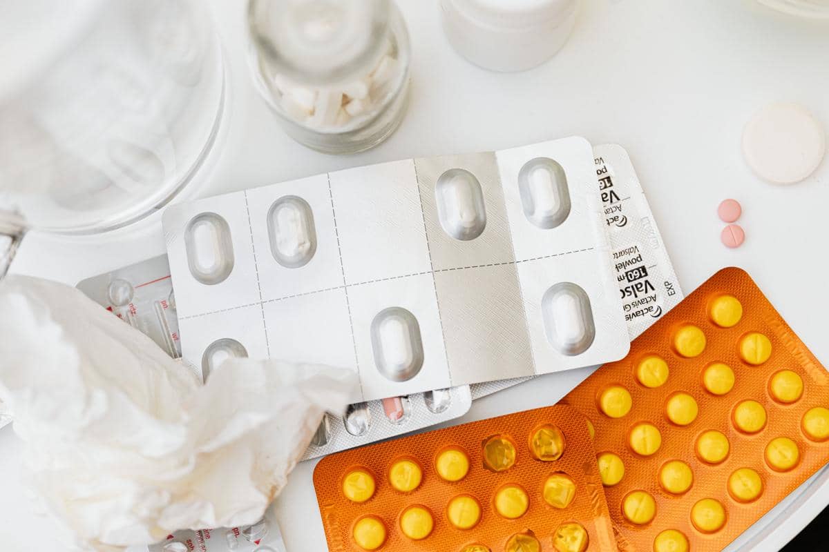 Beragam jenis obat dalam bentuk tablet dan kapsul dalam kemasan blister berwarna perak dan oranye di atas meja putih.