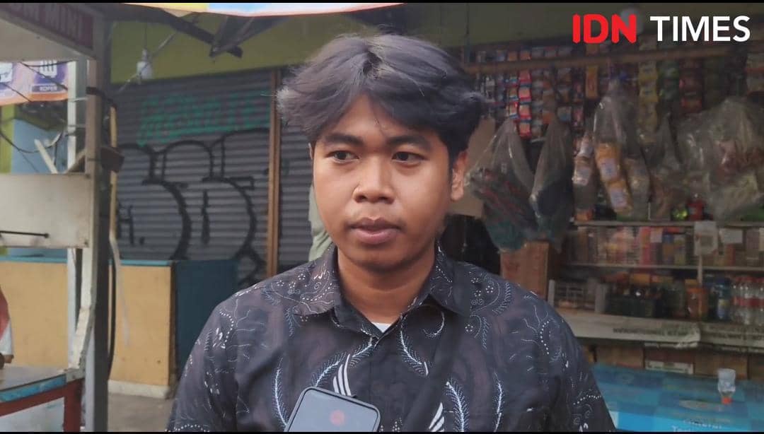 Seorang pria berambut hitam mengenakan kemeja bermotif gelap berdiri di depan kios kecil dengan latar barang dagangan dan pintu bergulung.