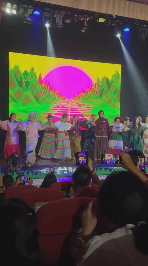 Anak-anak mengenakan kostum warna-warni menari di atas panggung dengan latar layar digital cerah di acara wisuda TK.