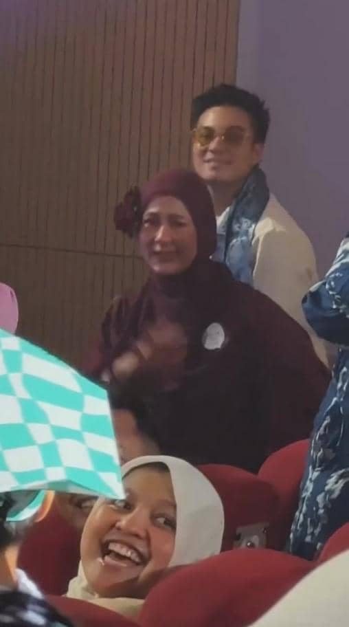 Seorang pria dan wanita tersenyum di antara penonton acara wisuda TK, duduk di kursi auditorium dengan suasana hangat dan bahagia.