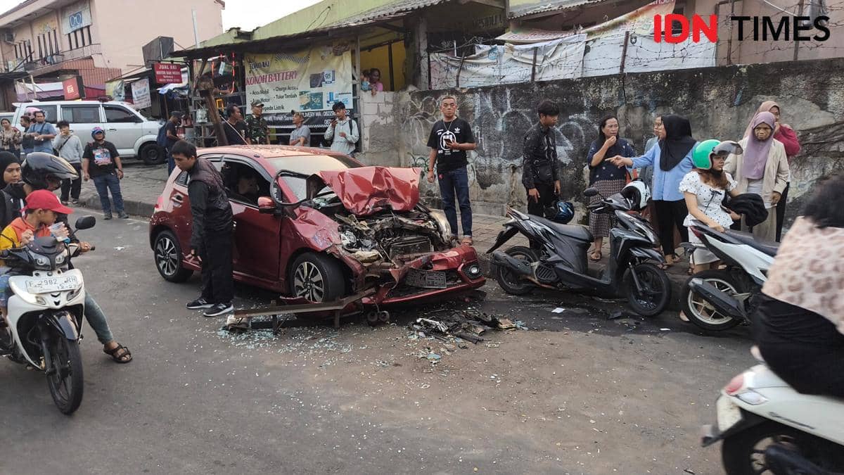 Mobil Brio merah rusak parah di bagian depan setelah kecelakaan di Bogor, dikelilingi warga dan pengendara motor yang menyaksikan.