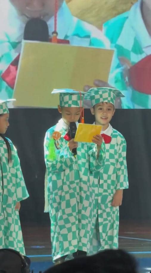 Anak-anak TK mengenakan toga bermotif kotak hijau putih sedang tampil di panggung wisuda sambil memegang kertas dan mikrofon.
