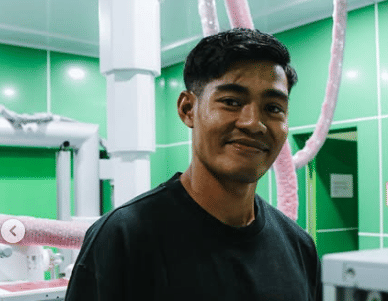 Seorang pria mengenakan kaus hitam tersenyum di dalam ruangan berwarna hijau dengan peralatan di sekitarnya.