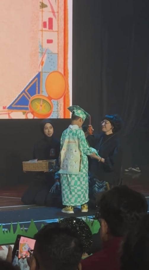 Seorang anak mengenakan toga hijau bermotif kotak sedang menerima penghargaan di atas panggung wisuda TK di depan para penonton.