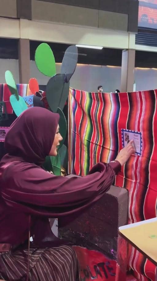 Seorang perempuan berhijab ungu tua mengenakan blus satin dan rok garis sedang menempelkan kertas pada dinding berwarna-warni di acara sekolah.
