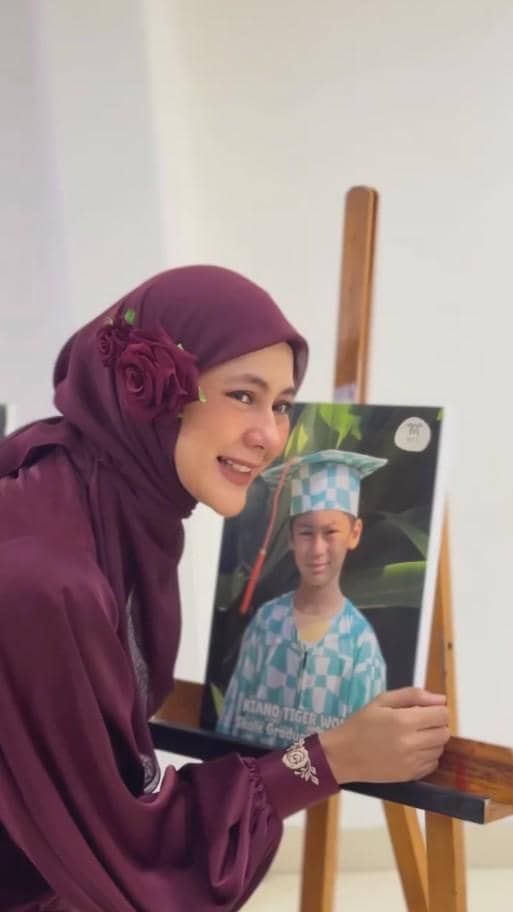 Seorang wanita berhijab ungu tersenyum sambil berpose di samping foto anak laki-laki mengenakan toga wisuda TK bermotif kotak biru putih.