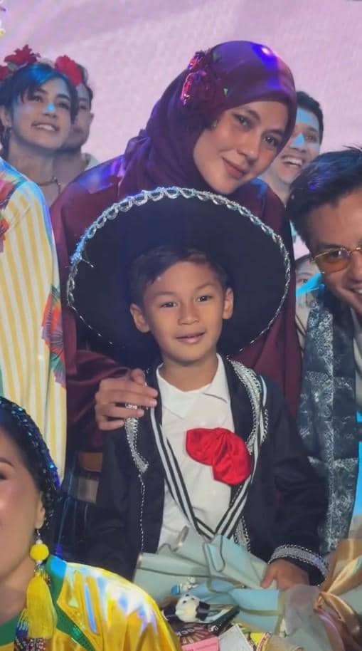 Seorang anak mengenakan kostum bertema Meksiko dengan topi sombrero hitam berpose bersama seorang wanita berhijab di acara wisuda TK.