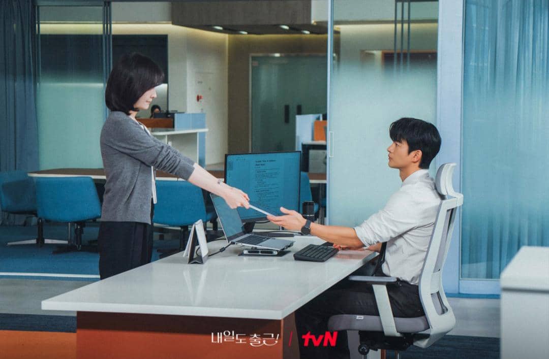 Seorang wanita menyerahkan dokumen kepada pria yang duduk di meja kantor modern dengan komputer dan suasana kerja profesional.