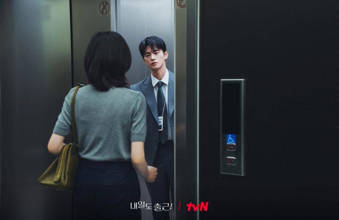 Seorang pria berpakaian jas berdiri di dalam lift menatap seorang wanita yang berdiri di luar lift dalam suasana kantor modern.