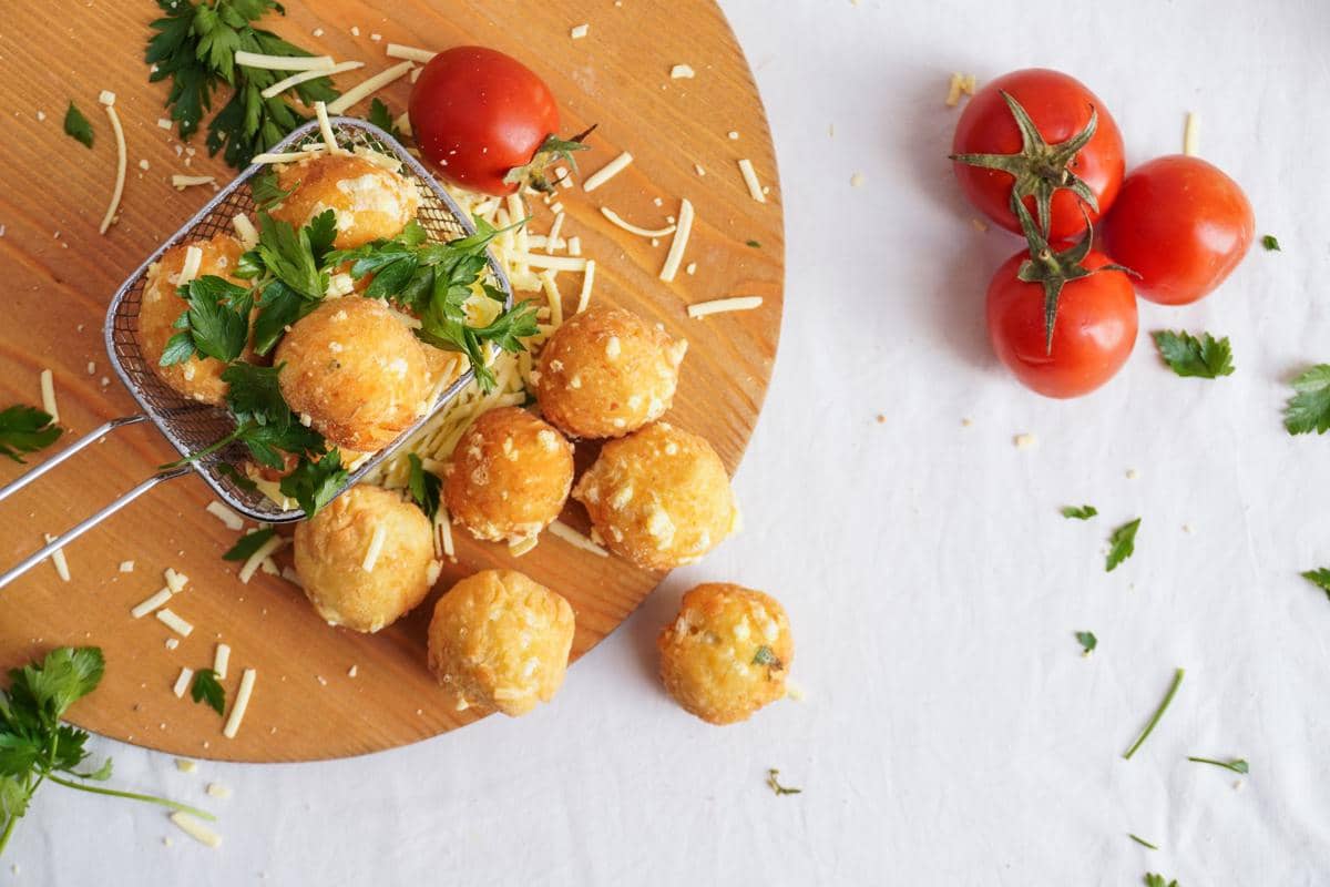 Beberapa cheese balls rendah kalori disajikan di atas talenan kayu dengan taburan keju parut, daun peterseli, dan tomat segar.