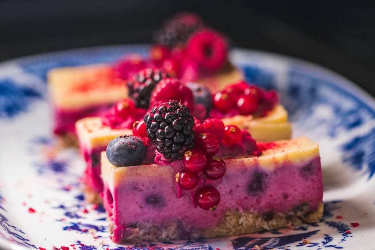 Potongan kue mousse berry berlapis warna ungu dan kuning dengan topping buah segar seperti blackberry, blueberry, dan red currant di atas piring bermotif biru.