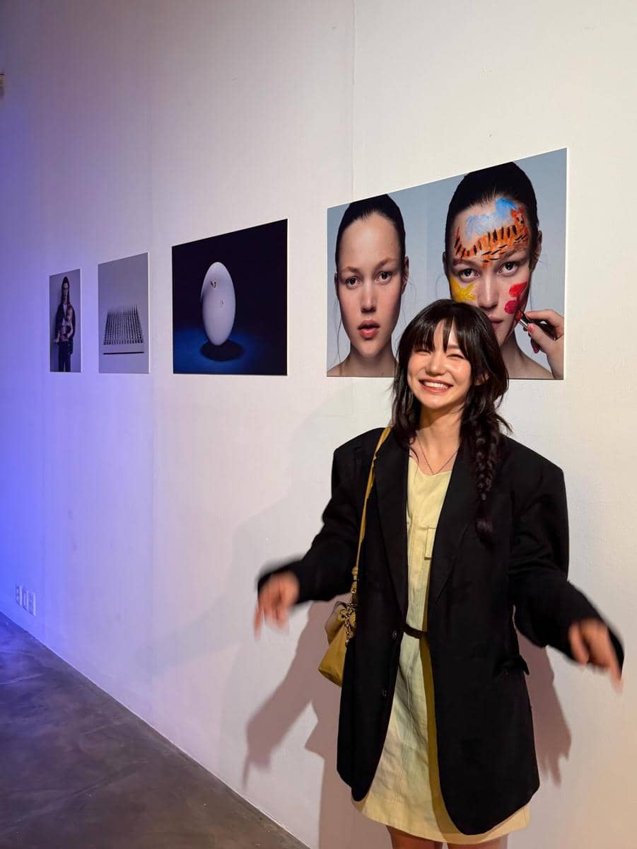 Seorang wanita tersenyum mengenakan blazer hitam dan gaun krem berdiri di depan dinding galeri dengan beberapa karya foto artistik.