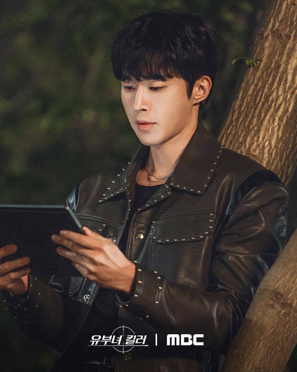 Seorang pria mengenakan jaket kulit hitam sedang memegang tablet di luar ruangan pada malam hari, dengan latar pepohonan.