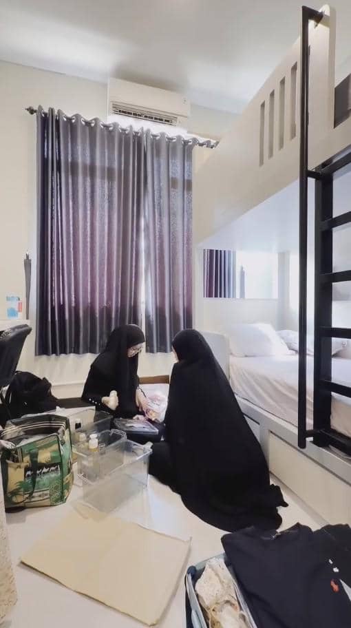Dua perempuan berhijab hitam sedang menata barang di kamar dengan tempat tidur tingkat dan koper terbuka di lantai.