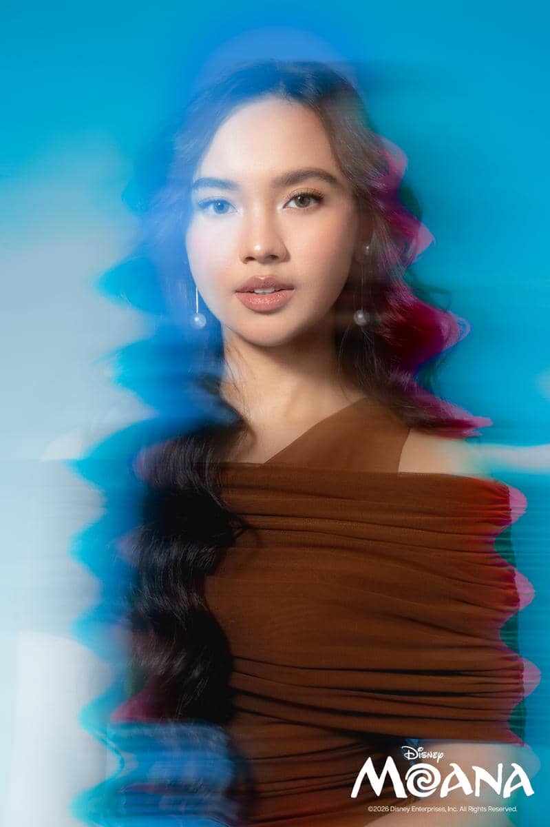 Seorang perempuan berambut panjang bergelombang mengenakan gaun cokelat dengan latar biru dan logo film Moana di sudut gambar.