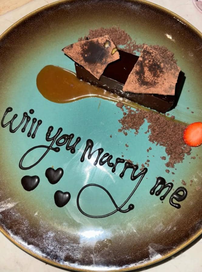 Sepiring dessert cokelat dengan tulisan 'Will you marry me' dari saus cokelat, dihiasi potongan stroberi dan saus karamel.