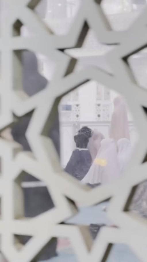 Seorang anak mengenakan pakaian muslim bersama beberapa orang di dalam ruangan berpola islami yang terlihat melalui ukiran dinding.