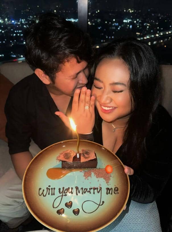 Seorang pria melamar kekasihnya sambil memegang piring bertuliskan 'Will you marry me' dengan kue cokelat dan lilin menyala di atasnya.