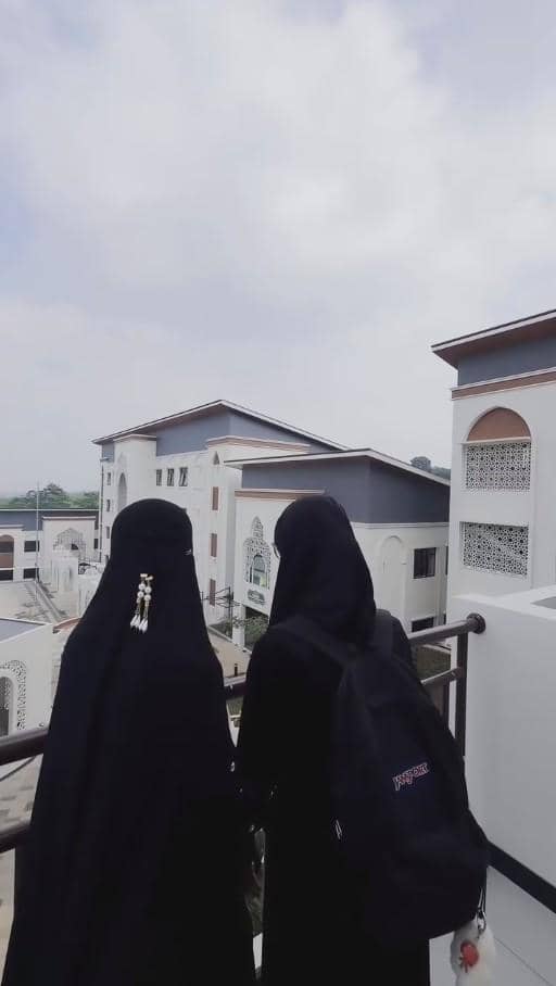 Dua perempuan berpakaian hitam lengkap dengan cadar berdiri di balkon menghadap bangunan pesantren berarsitektur modern.