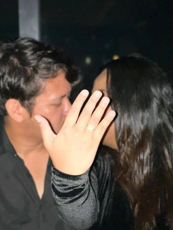 Seorang wanita memperlihatkan cincin di jarinya sambil berpose mesra dengan seorang pria dalam suasana malam yang redup.