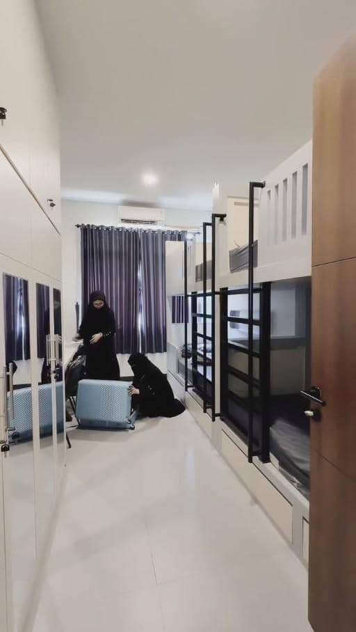 Dua perempuan berpakaian hitam sedang menata koper biru di kamar asrama dengan tempat tidur susun dan tirai berwarna ungu.