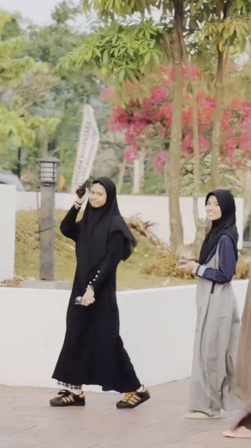 Seorang perempuan mengenakan gamis hitam dan kerudung hitam berjalan bersama seorang gadis muda di area terbuka dengan pepohonan dan bunga berwarna cerah.