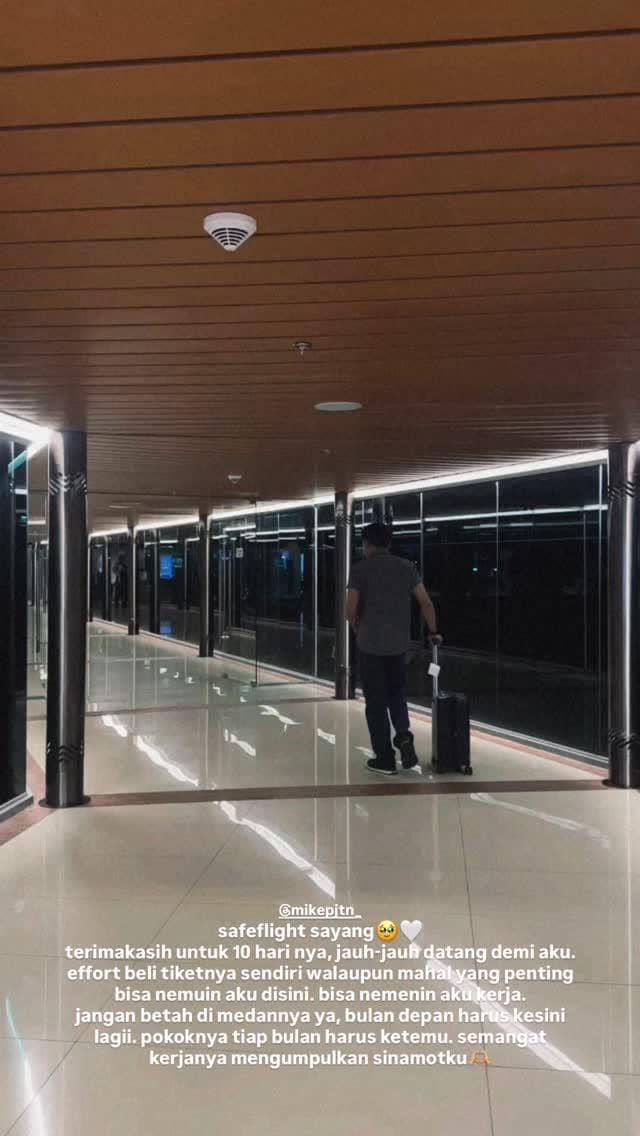 Seorang pria berjalan di lorong bandara dengan membawa koper kecil, tampak suasana malam dengan pencahayaan modern di sekitarnya.