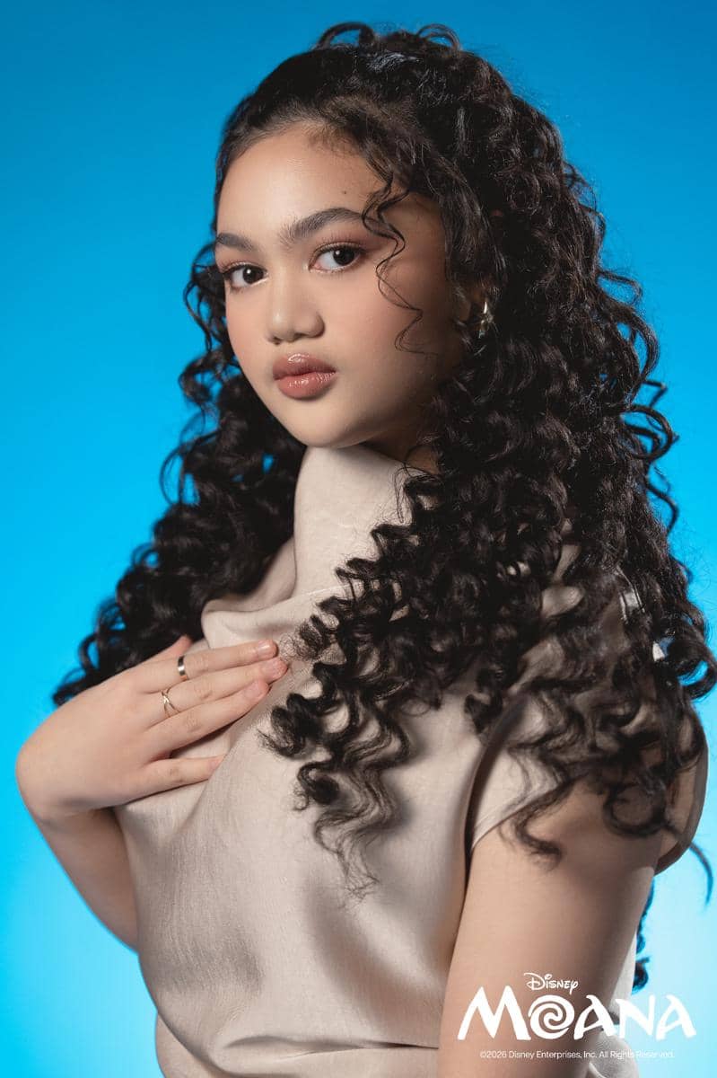 Seorang perempuan berambut ikal panjang mengenakan pakaian berwarna krem berpose di depan latar biru dengan logo Disney Moana di sudut gambar.