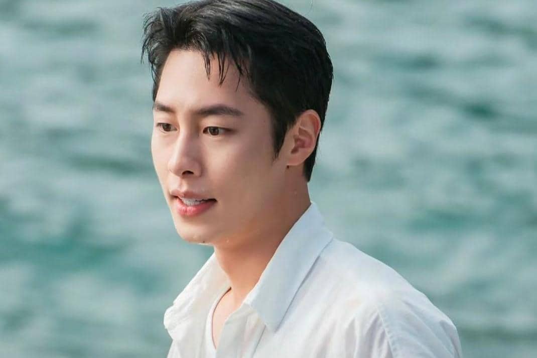 Seorang pria mengenakan kemeja putih berdiri di tepi laut dengan latar air berwarna biru kehijauan dalam adegan drama Doctor On The Edge.