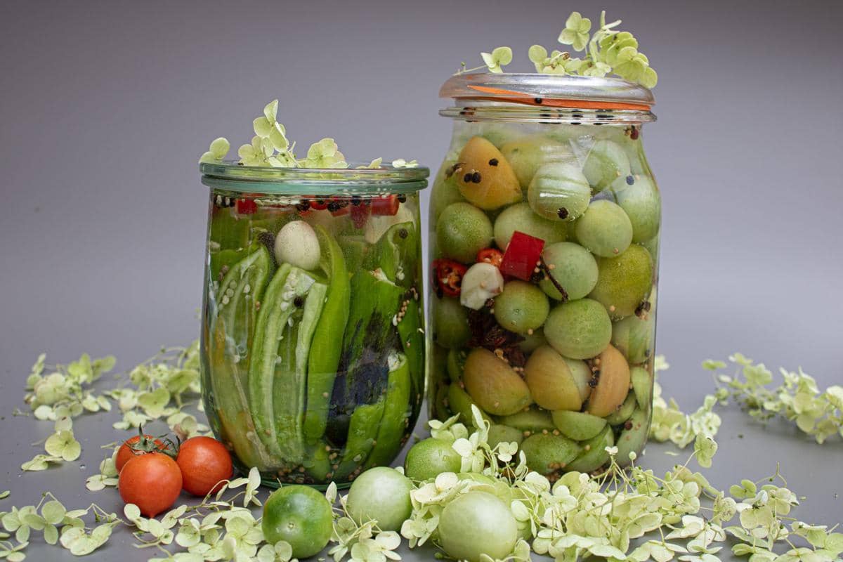 Dua toples kaca berisi asinan ceri hijau dan sayuran dengan bumbu rempah, dikelilingi buah tomat dan daun hijau di atas meja abu-abu.